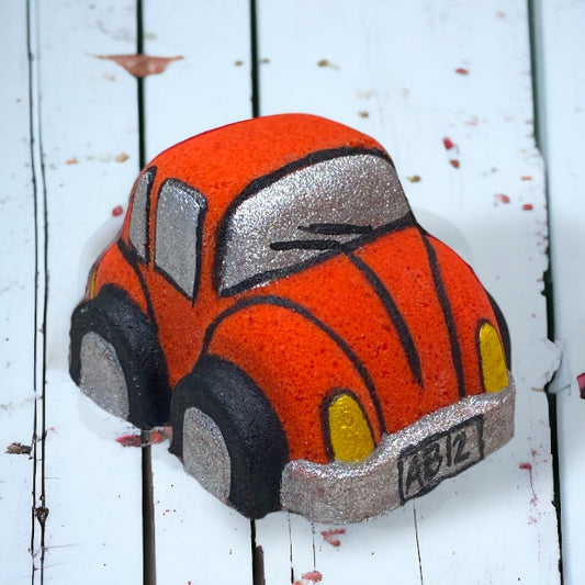 A red orange Volkswagen Bug car-shaped bath bomb with black and yellow accents on a wooden surface with water droplets around it.