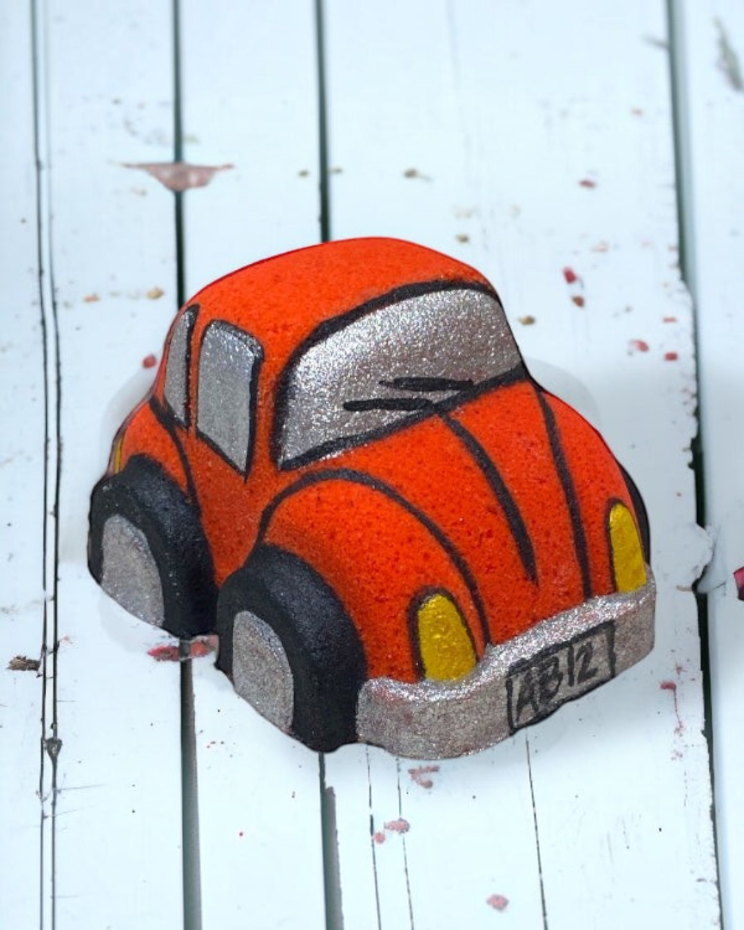 A red orange Volkswagen Bug car-shaped bath bomb with black and yellow accents on a wooden surface with water droplets around it.
