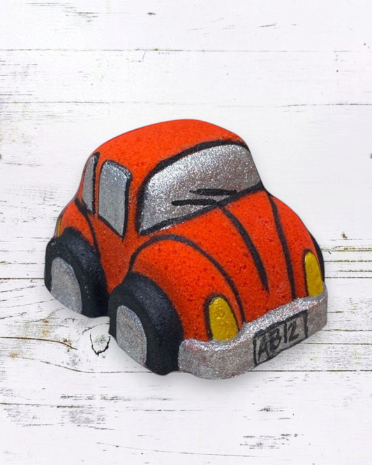 A red orange Volkswagen Bug car-shaped bath bomb with black and yellow accents on a wooden surface with water droplets around it.