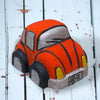 A red orange Volkswagen Bug car-shaped bath bomb with black and yellow accents on a wooden surface with water droplets around it.
