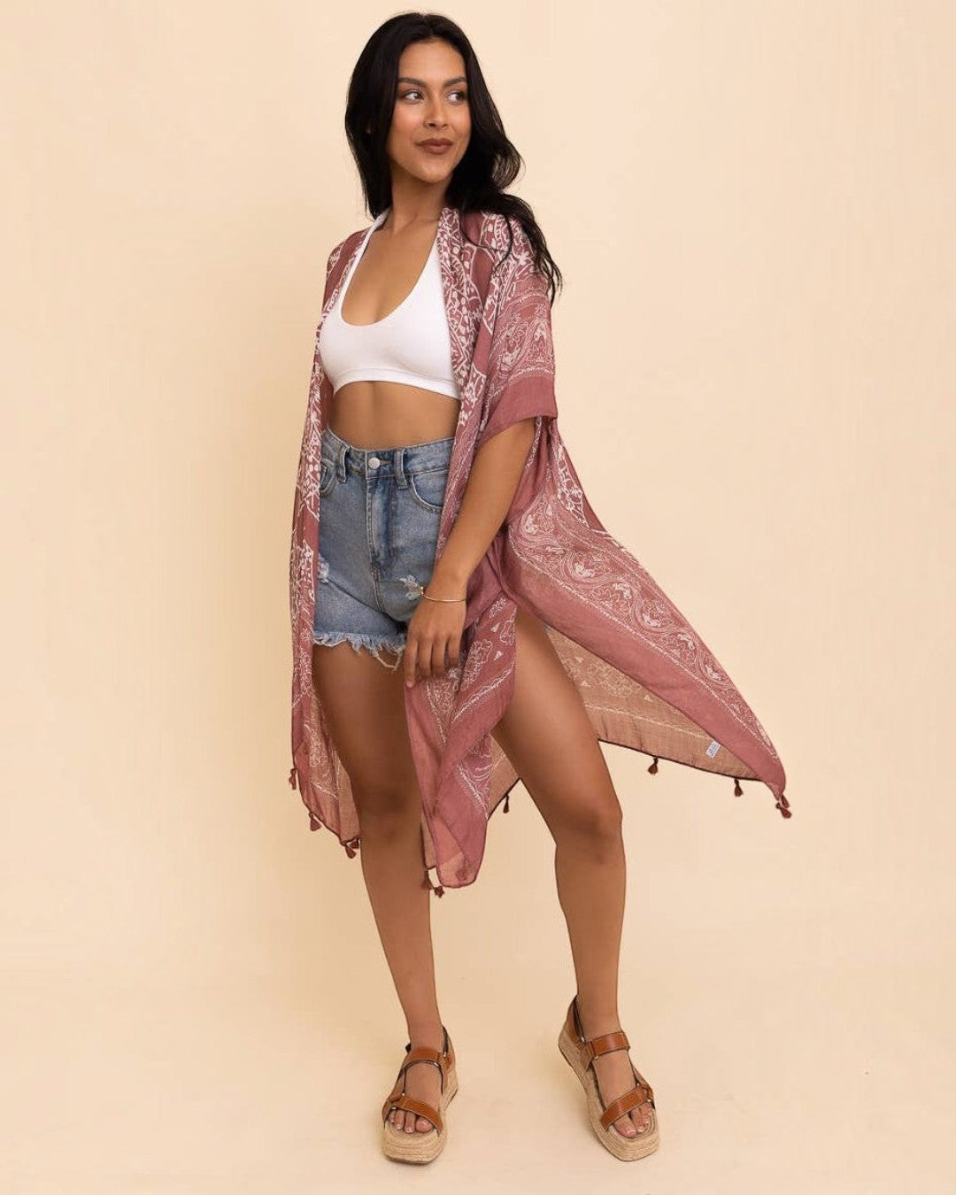 Woman wearing a pink lace kimono over a white top and denim shorts on a beige background