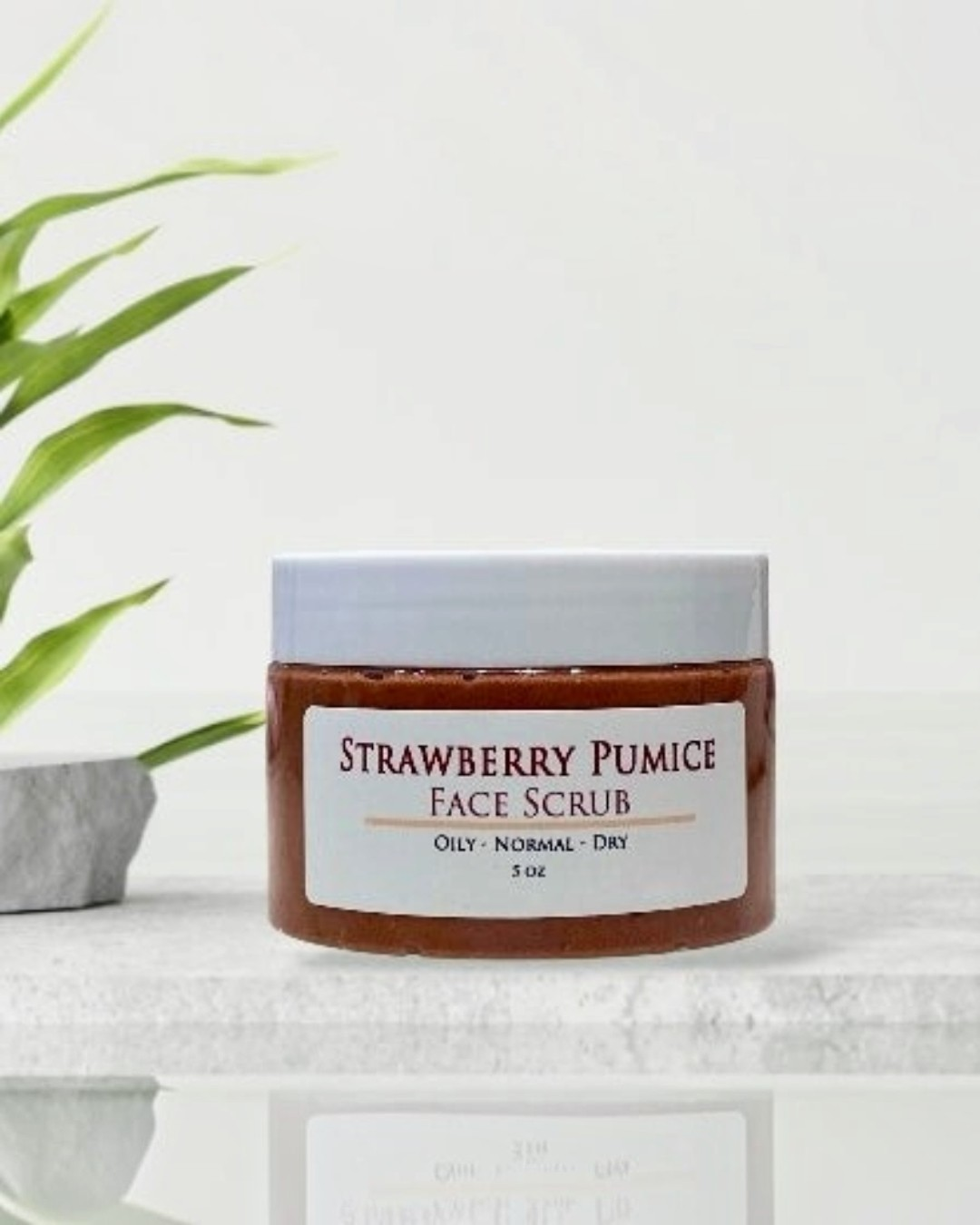 Jar of Strawberry Pumice Face Scrub on a marble surface with a plant in the background