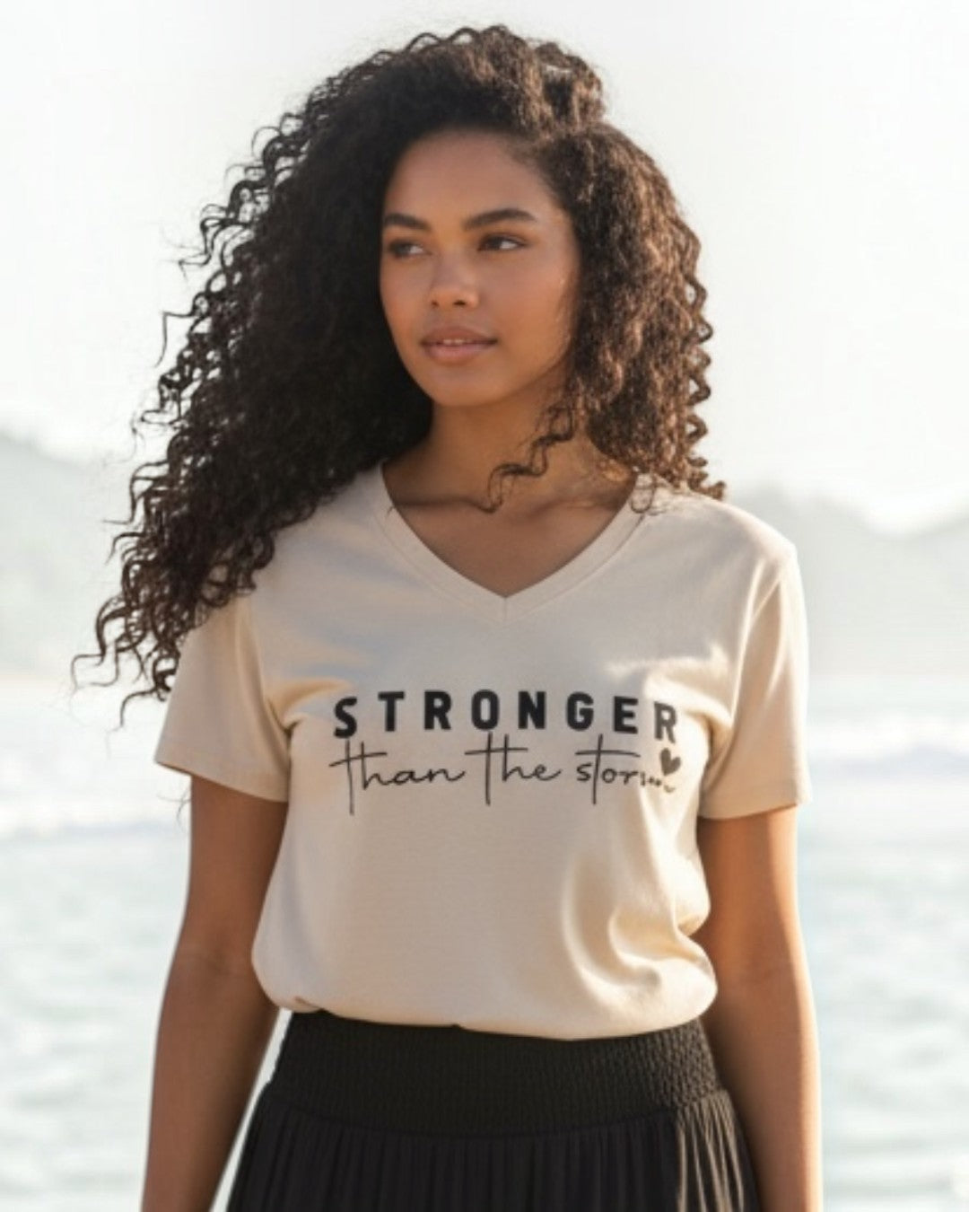 Woman standing on a beach wearing a beige t-shirt with text and a black skirt.