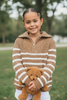 Young girl wearing a brown and white striped sweater holding a teddy bear outdoors.