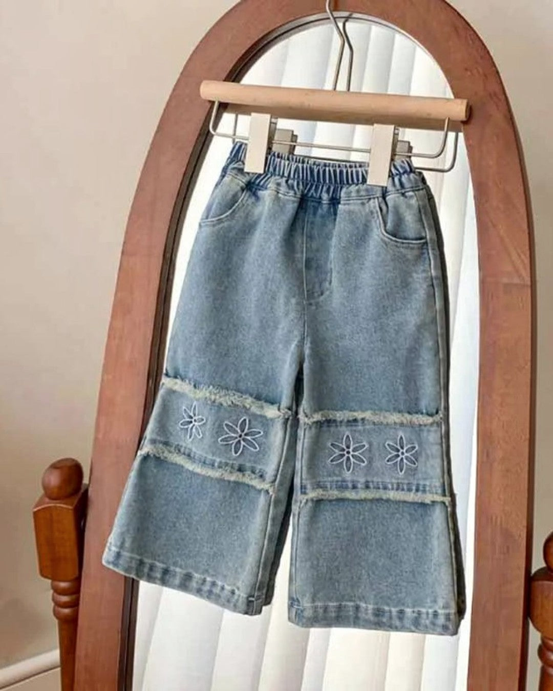 Denim pants with floral embroidery on a hanger in front of a mirror.
