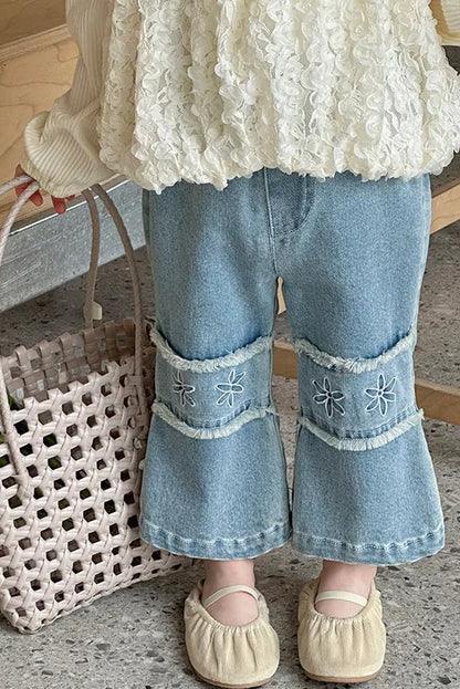 Person wearing a textured white top and blue jeans with floral embroidery, holding a woven bag with flowers.