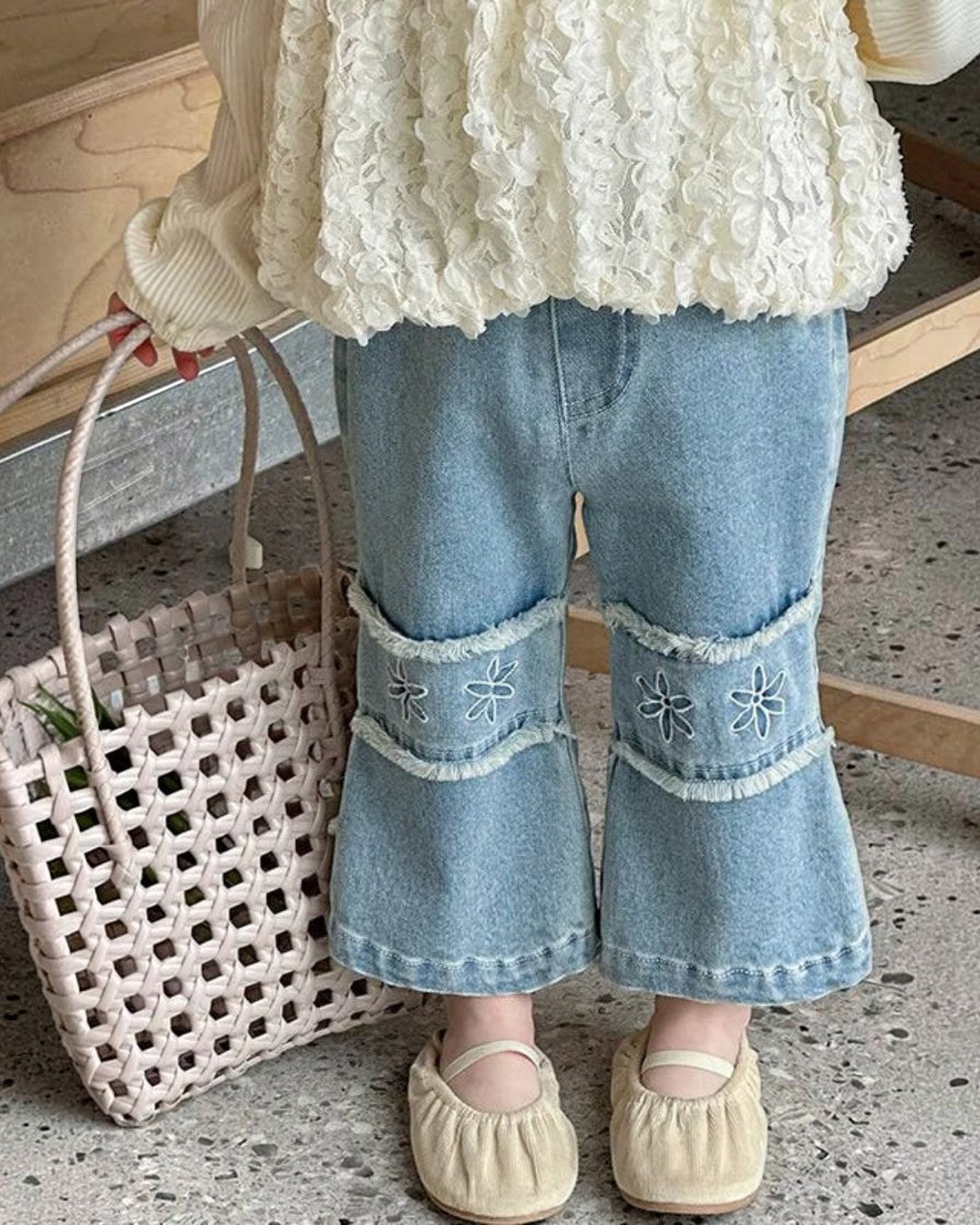 Person wearing a textured white top and blue jeans with floral embroidery, holding a woven bag with flowers.