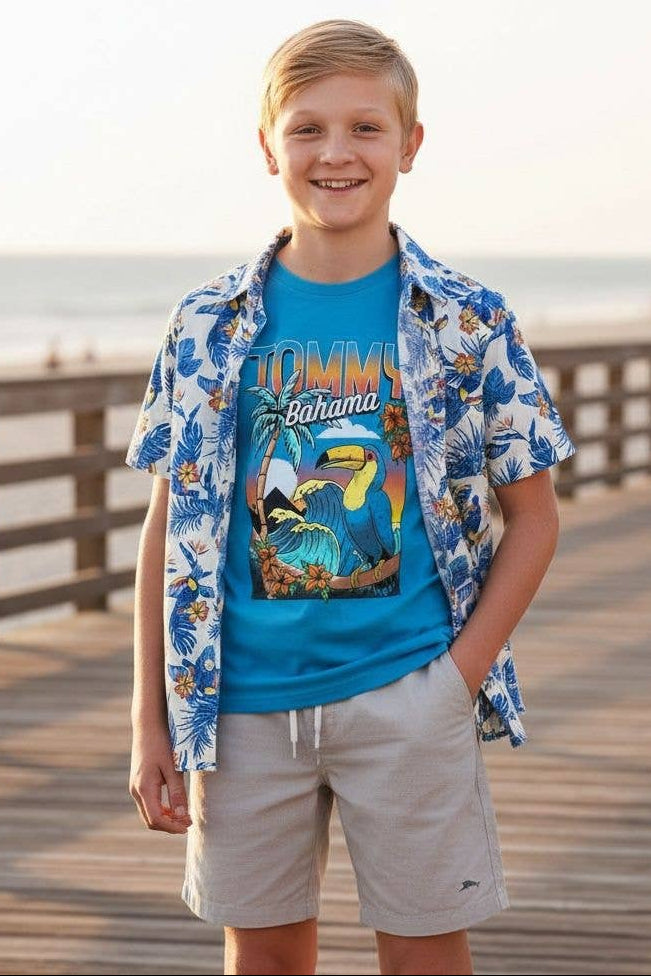 Young boy in Tommy Bahama shirt and shorts standing on a wooden boardwalk by the ocean.