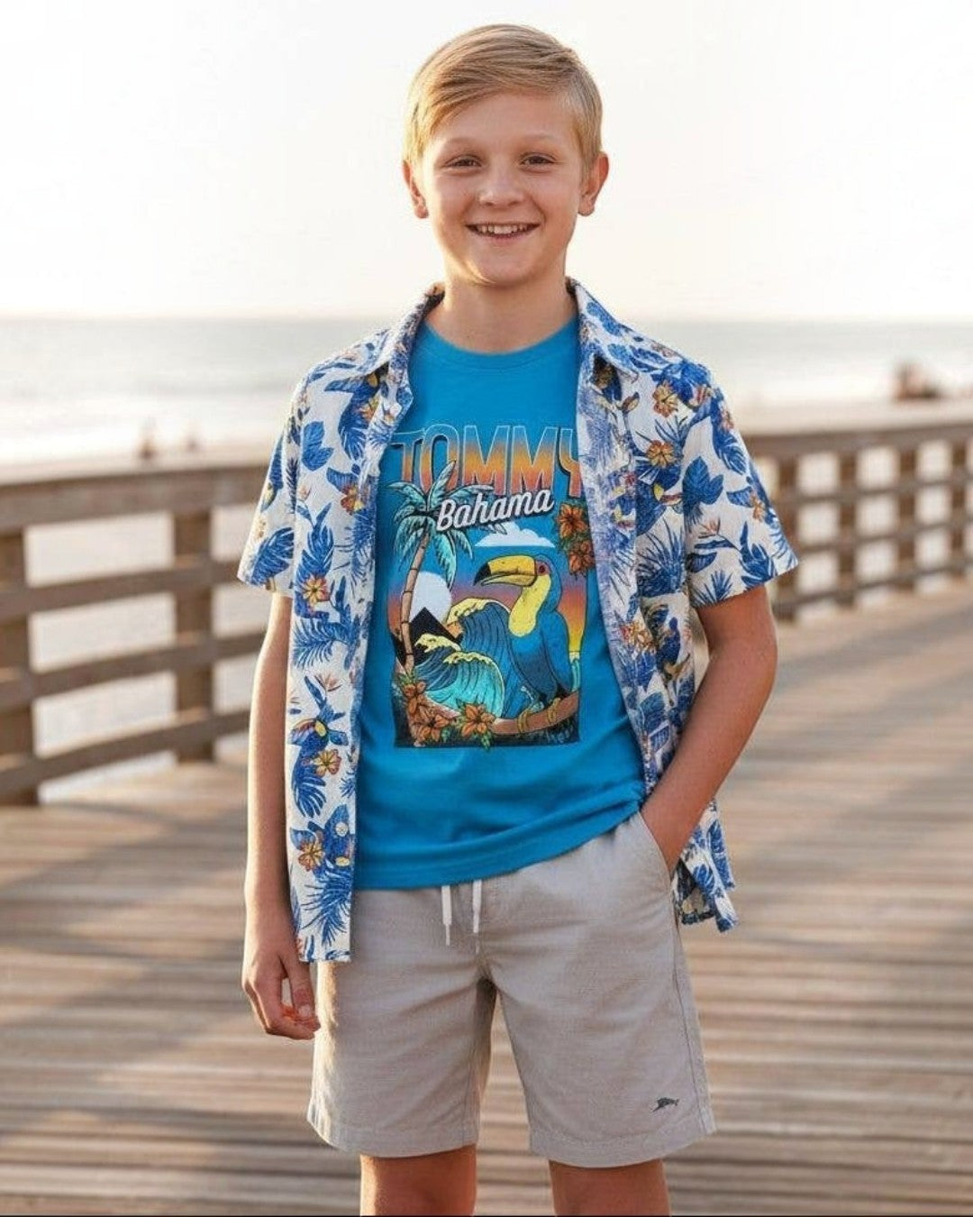 Young boy in Tommy Bahama shirt and shorts standing on a wooden boardwalk by the ocean.