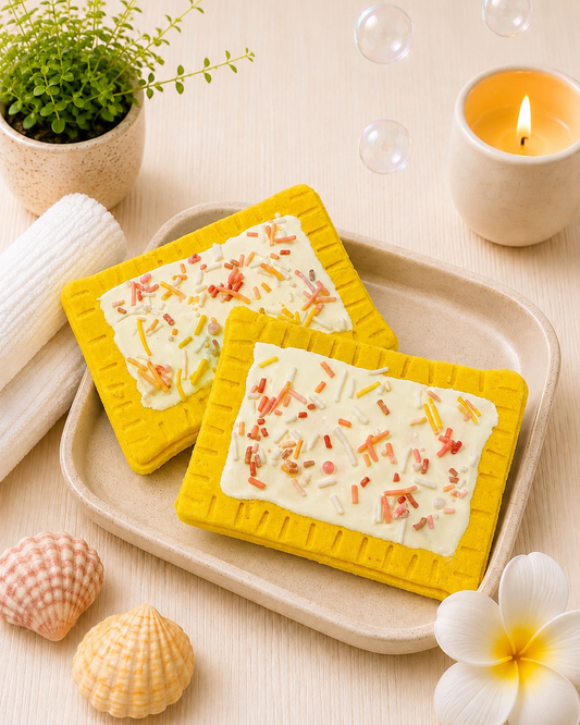 Two rectangular soap bars with colorful toppings on yellow soap dishes, placed on a beige tray with a candle, plant, and flowers in the background.