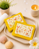 Two rectangular soap bars with colorful toppings on yellow soap dishes, placed on a beige tray with a candle, plant, and flowers in the background.