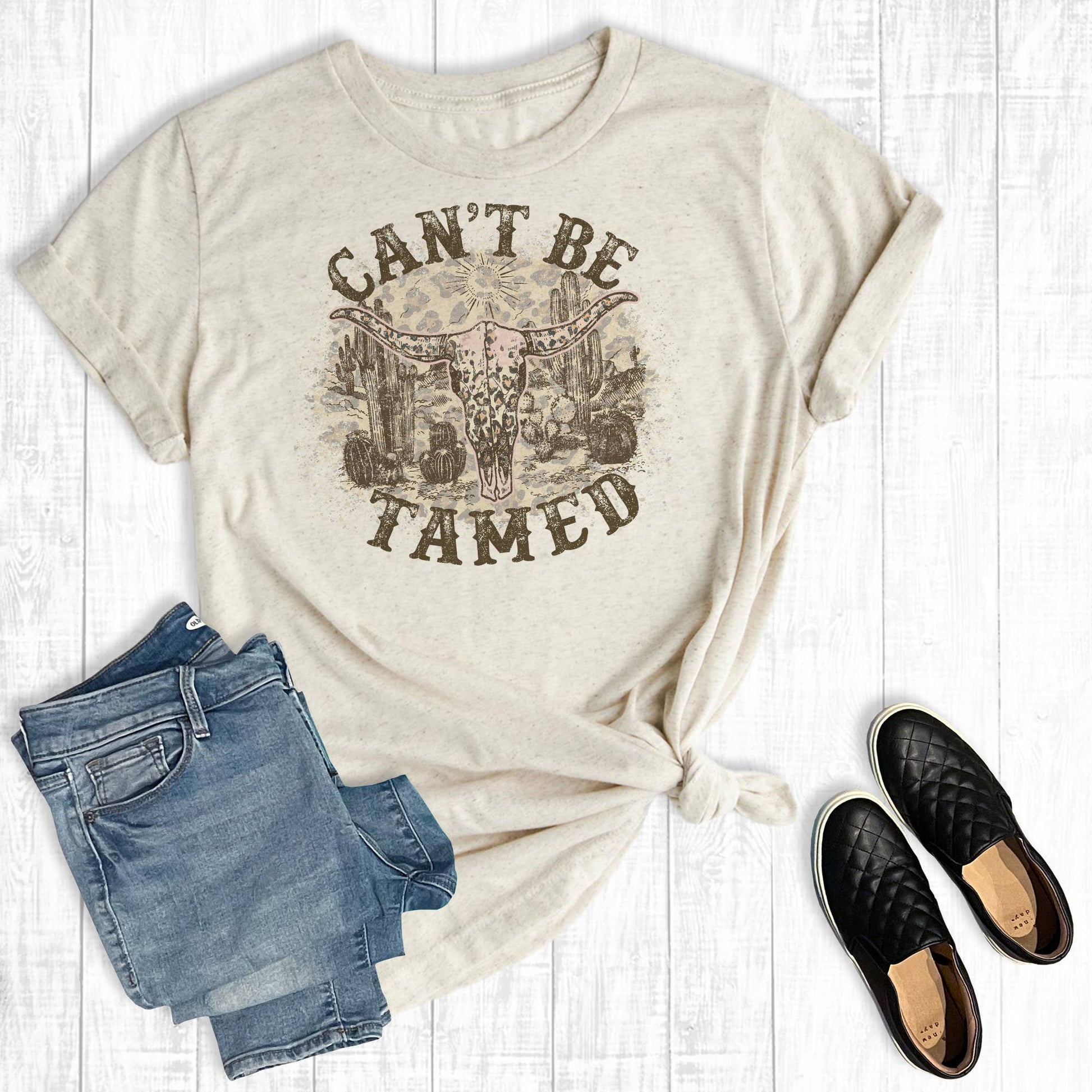 Beige t-shirt with graphic design, denim shorts, and black shoes on a light wooden surface