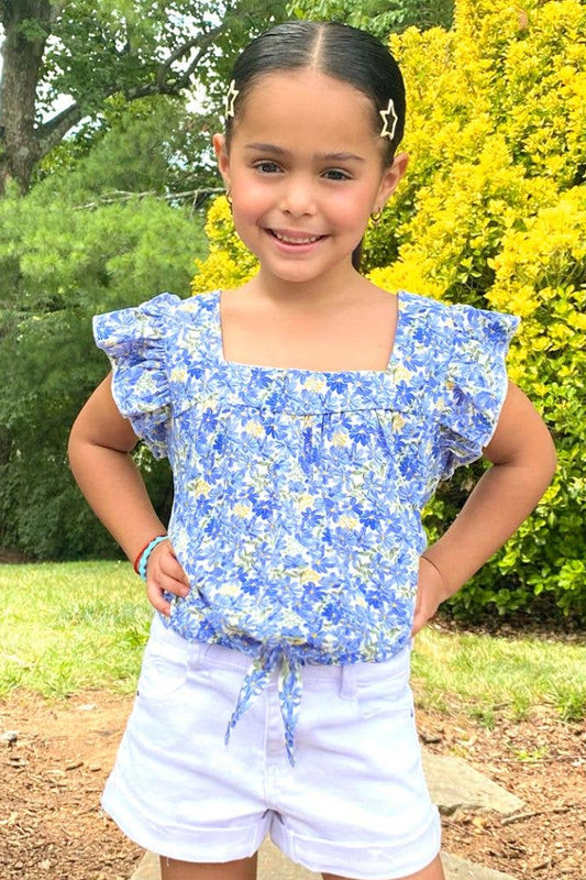 Young girl wearing a blue floral top and white shorts standing outdoors with greenery in the background