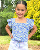 Young girl wearing a blue floral top and white shorts standing outdoors with greenery in the background