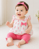 Baby girl in a white outfit with pink accents sitting on a light-colored floor.