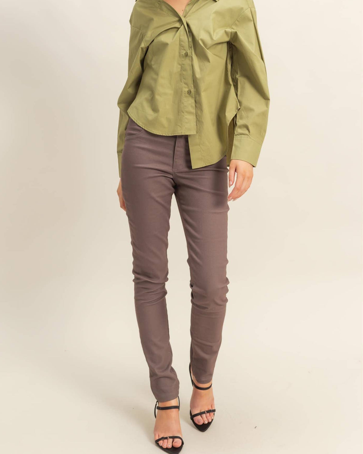 A woman modeling a sage green button-down shirt and taupe slim-fit pants with black high-heeled sandals, standing against a plain light beige studio background.