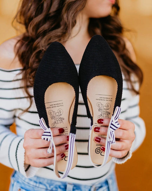 Black flats with decorative bows held by a person wearing a striped shirt.