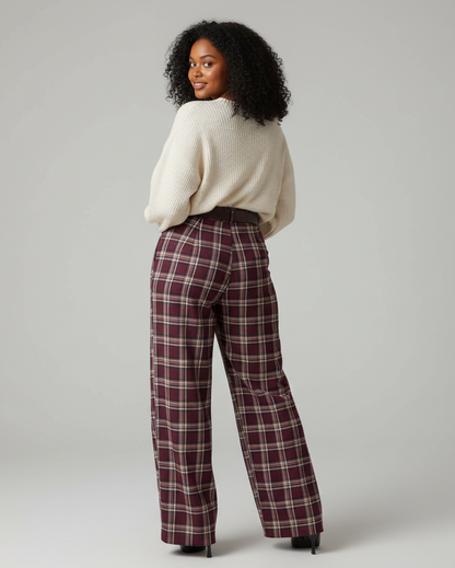 A woman stands with her back turned wearing a cream knit sweater and high-waisted, wide-leg plaid trousers in burgundy, beige, and white. She is photographed against a neutral light gray studio background.