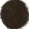 A pile of dark, finely ground loose-leaf tea or herbal blend filling a light-colored container.