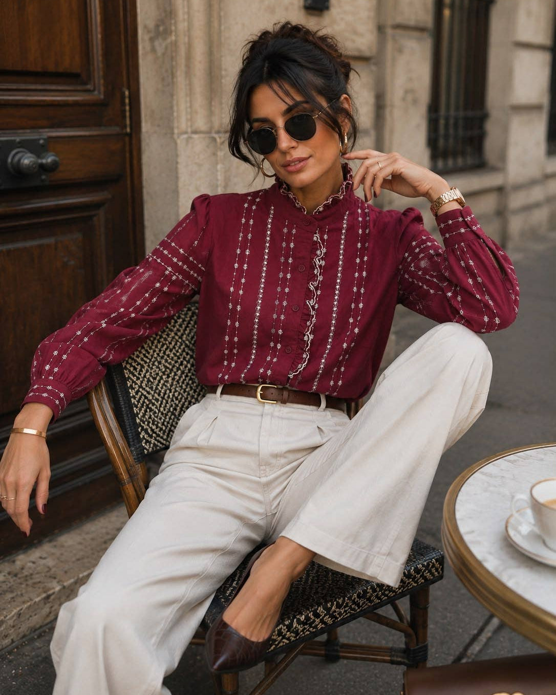 A woman wearing a burgundy long-sleeve blouse with vertical white embroidered patterns, paired with white wide-leg trousers and brown pointed-toe flats, sitting on a cafe chair.