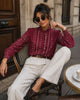 A woman wearing a burgundy long-sleeve blouse with vertical white embroidered patterns, paired with white wide-leg trousers and brown pointed-toe flats, sitting on a cafe chair.
