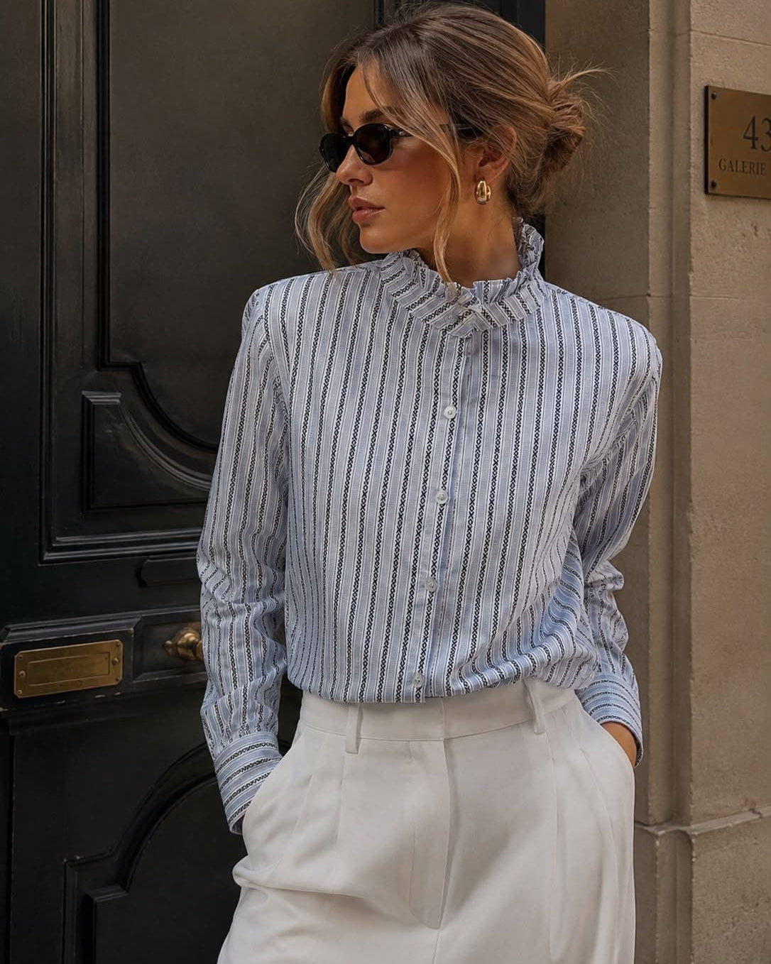 A woman wearing a light blue and white vertically striped long-sleeve blouse with a ruffled high neck, paired with white trousers, standing in front of a dark wooden door.