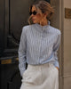 A woman wearing a light blue and white vertically striped long-sleeve blouse with a ruffled high neck, paired with white trousers, standing in front of a dark wooden door.