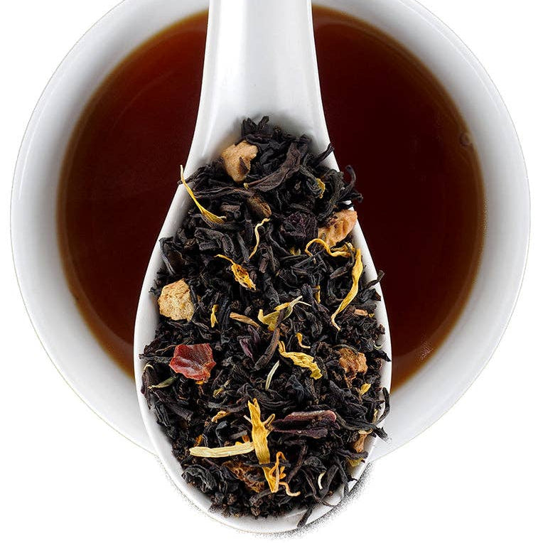 A white ceramic spoon filled with loose-leaf tea is shown over a white cup of dark tea on a white background. The tea blend contains dark black tea leaves with dried botanicals.