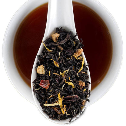 A white ceramic spoon filled with loose-leaf tea is shown over a white cup of dark tea on a white background. The tea blend contains dark black tea leaves with dried botanicals.