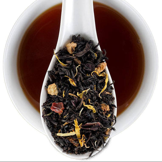 A white ceramic spoon filled with loose-leaf tea is shown over a white cup of dark tea on a white background. The tea blend contains dark black tea leaves with dried botanicals.