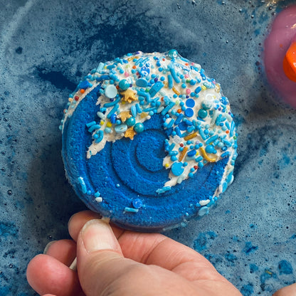 Blue bath bomb with colorful sprinkles held by a hand on a blue textured surface