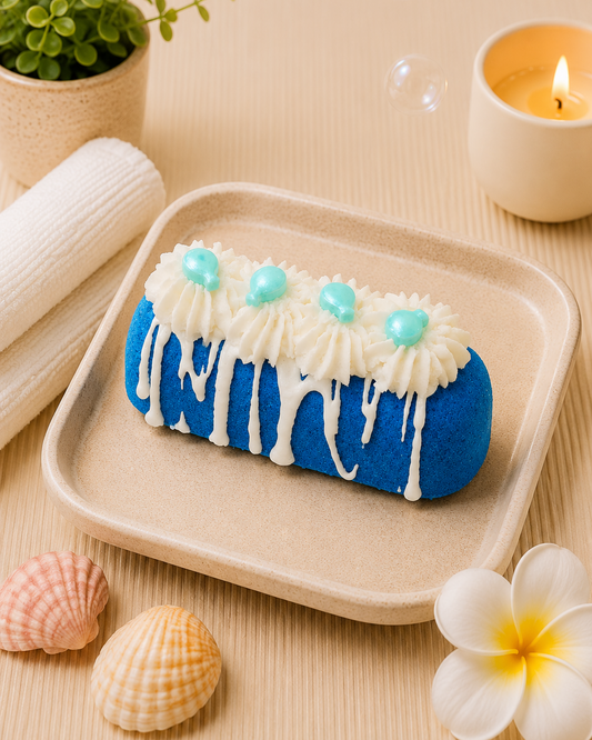 A blue and white decorated pastry on a beige tray, styled with seashells, a small plant, a lit candle, and a plumeria flower on a light beige surface.