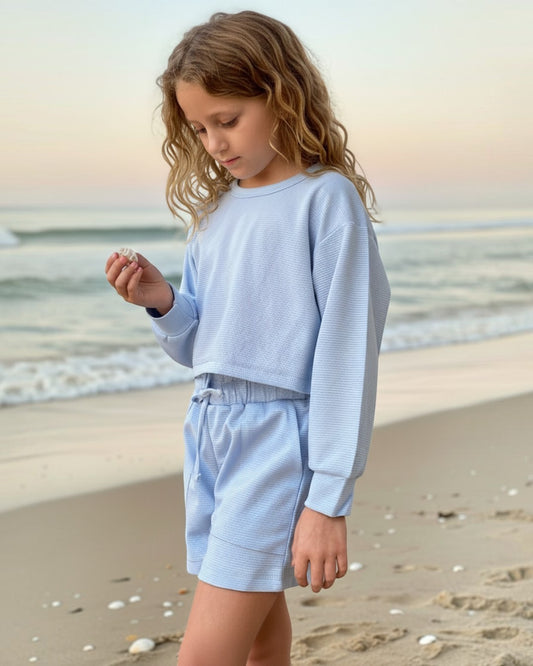 Girl wearing a Calla Lily Cosmetics + CL kids baby blue waffle knit lounge set with matching shorts on the beach.