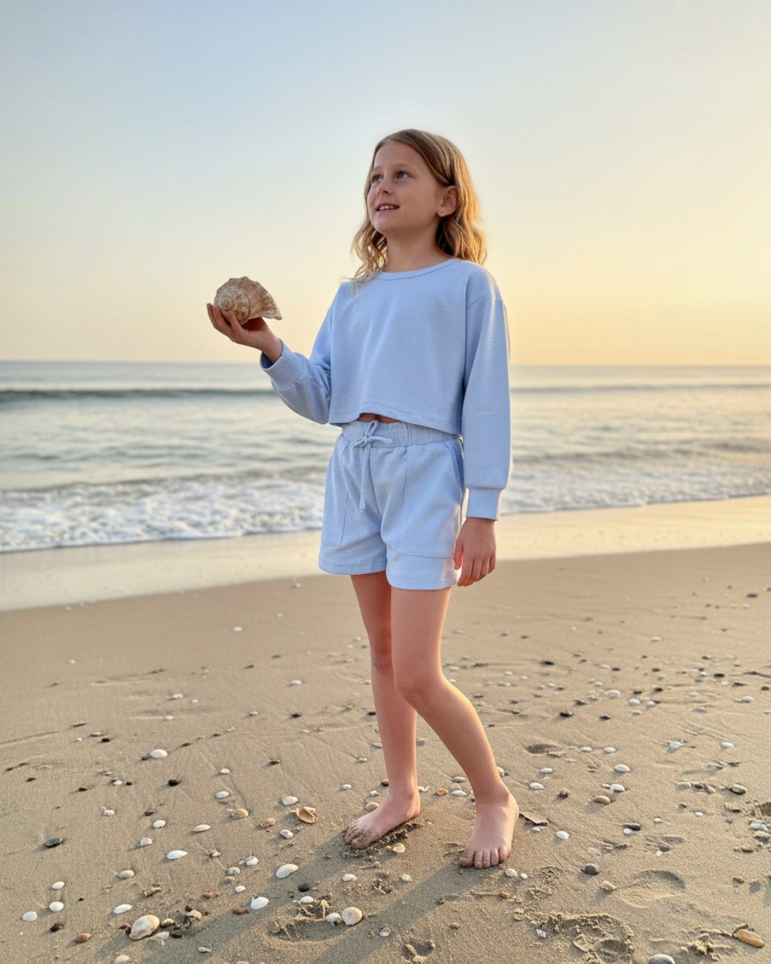 Girl wearing Calla Lily Cosmetics + CL kids baby blue waffle knit lounge set with shorts on a beach at sunset.