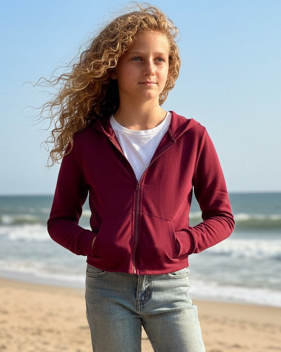 Calla Lily Cosmetics + CL kids burgundy French Terry crop zip-up hoodie worn by a girl on a beach boardwalk.