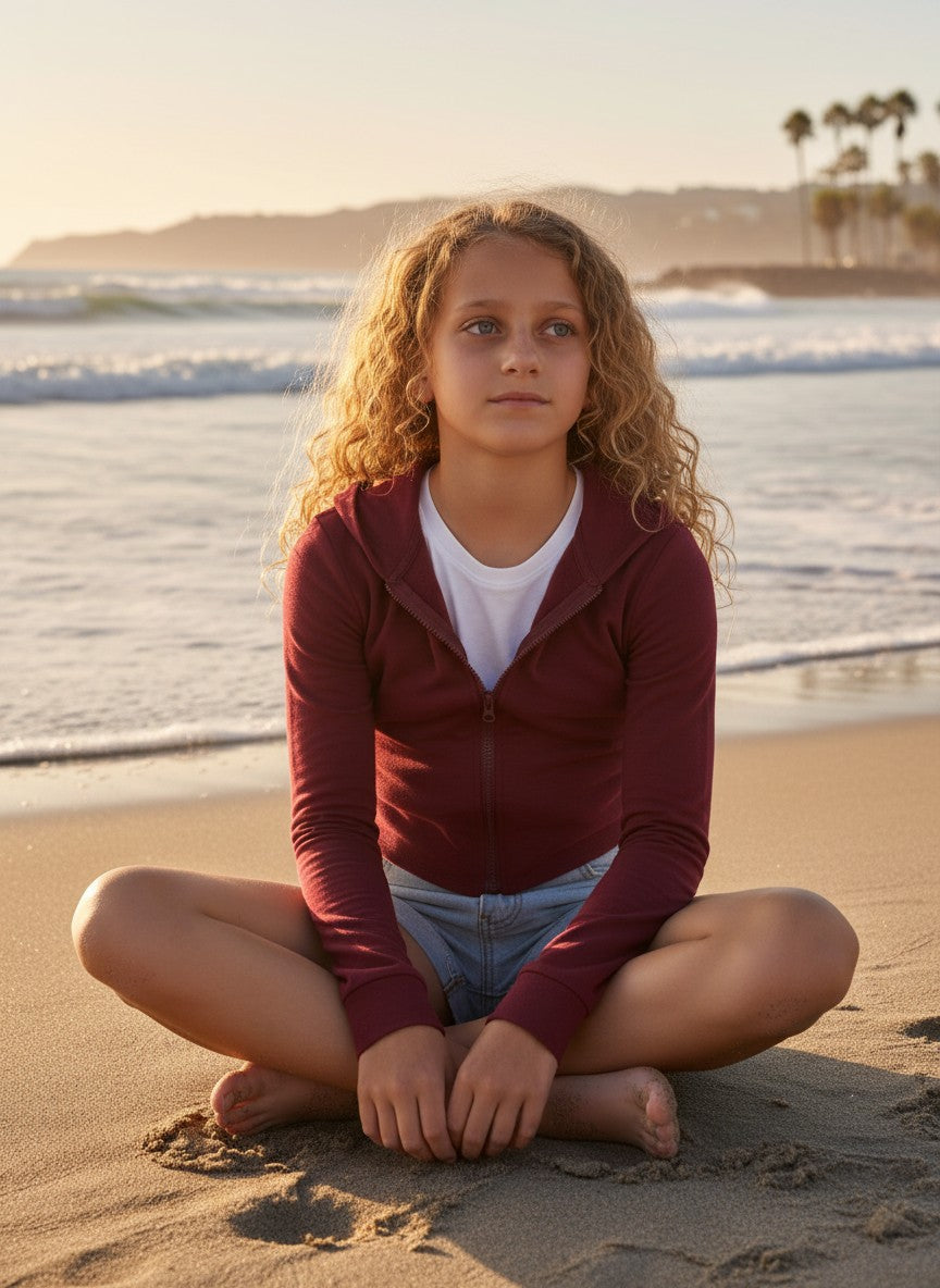 Model wearing a burgundy French Terry crop zip-up hoodie by Calla Lily Cosmetics + CL kids on a sunny beach.