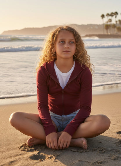 Model wearing a burgundy French Terry crop zip-up hoodie by Calla Lily Cosmetics + CL kids on a sunny beach.