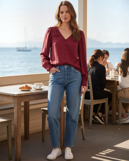 Model wearing a burgundy Blu Pepper satin V-neck blouse with long sleeves and blue jeans at a seaside cafe.