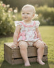 Baby wearing a Calla Lily Cosmetics + CL kids pastel floral bubble romper with pink ruffles, sitting on a wooden crate.