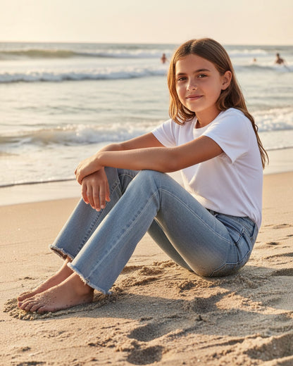 Girl wearing Calla Lily Cosmetics + CL kids light wash flare jeans with frayed hems, sitting on a sandy beach.