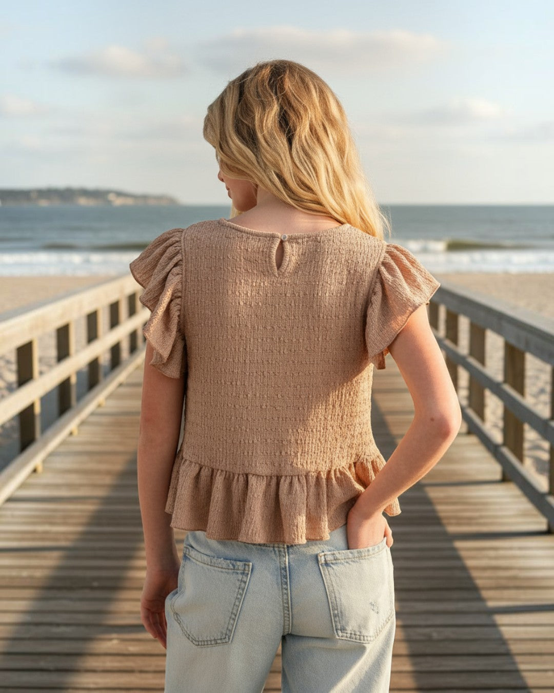 Girl wearing a beige textured knit top with ruffle sleeves by Calla Lily Cosmetics + CL kids on a beach boardwalk.