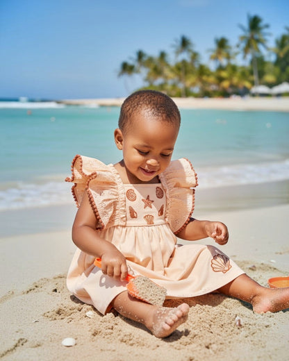 Baby girl wearing a Calla Lily Cosmetics + CL kids HoneyDrift dress with seashell embroidery on a sunny beach.