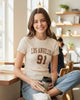 Woman wearing a Calla Lily Cosmetics + CL kids cream ribbed crew neck graphic tee with Los Angeles 91 print in a cafe