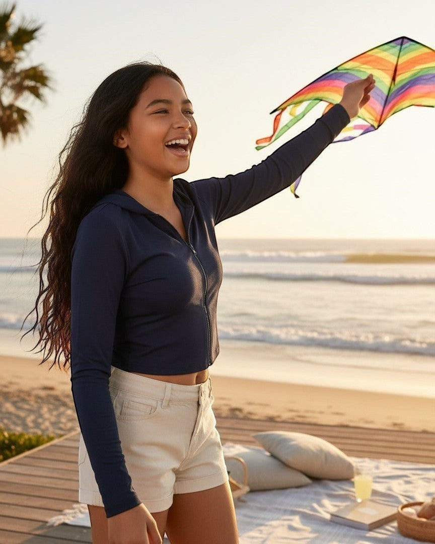 Model wearing Calla Lily Cosmetics + CL kids navy French Terry crop zip-up hoodie while flying a kite on the beach.