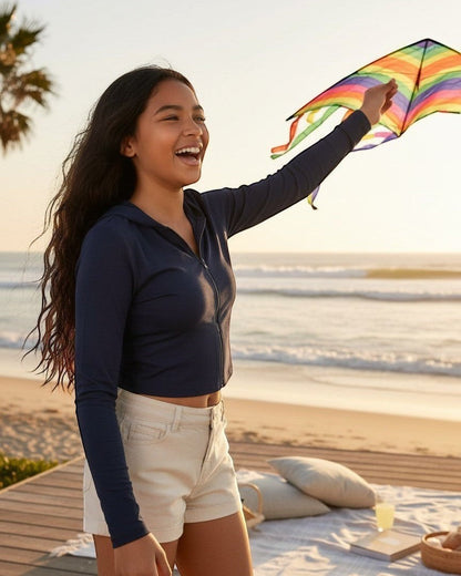 Model wearing Calla Lily Cosmetics + CL kids navy French Terry crop zip-up hoodie while flying a kite on the beach.