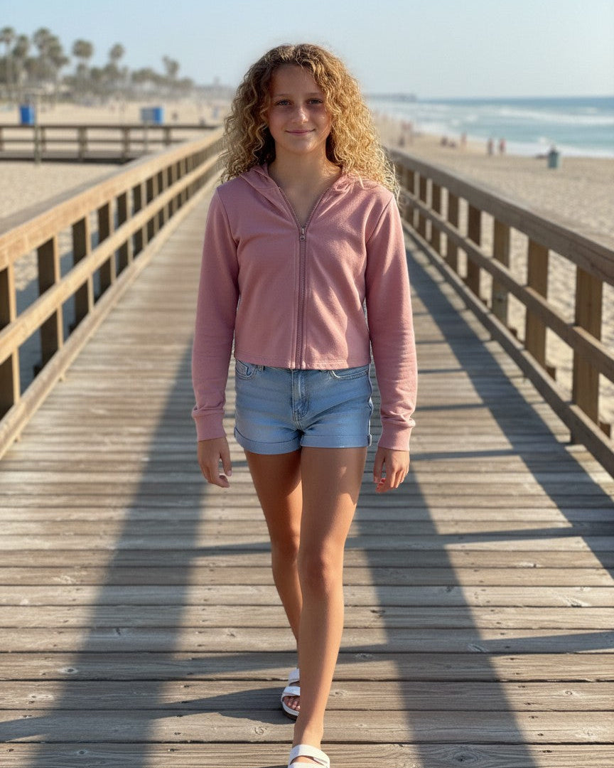 Model wearing a Calla Lily Cosmetics + CL kids pink French Terry crop zip-up hoodie on a wooden beach boardwalk.