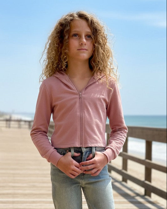 Model wearing a pink French Terry Crop Zip Up Hoodie by Calla Lily Cosmetics + CL kids on a sunny beach boardwalk.