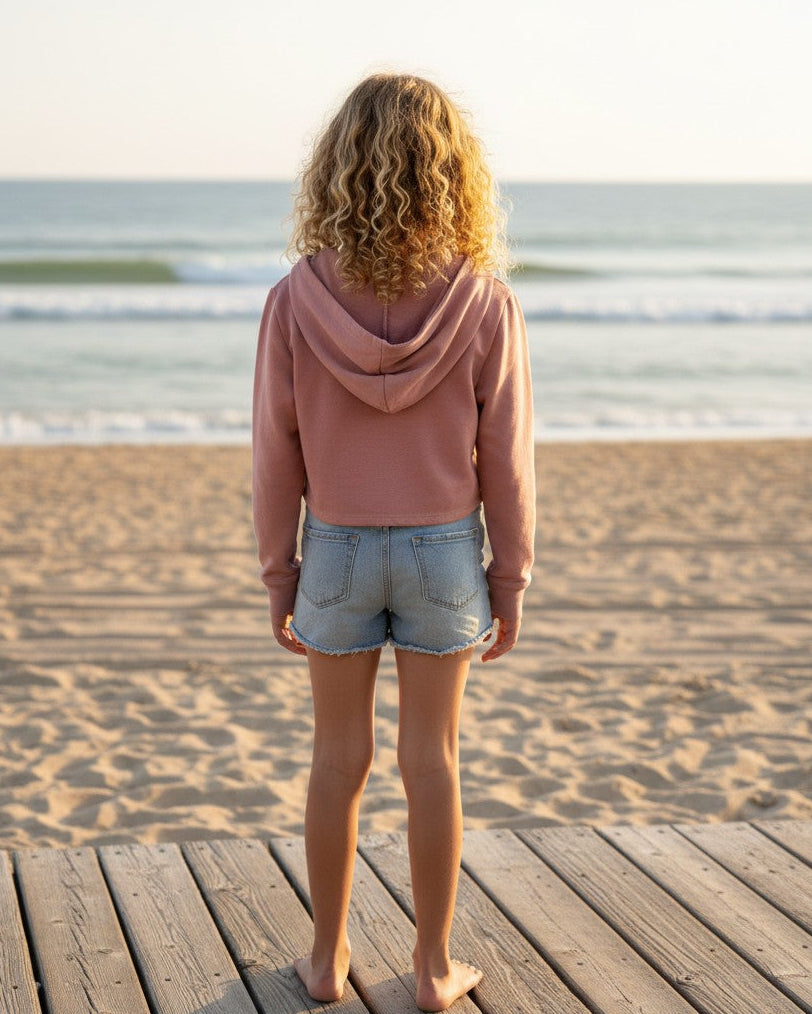 Calla Lily Cosmetics + CL kids pink French Terry crop hoodie worn by a girl with curly hair on a beach boardwalk.