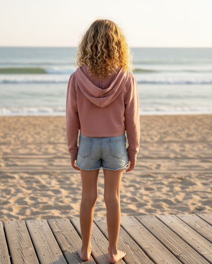 Calla Lily Cosmetics + CL kids pink French Terry crop hoodie worn by a girl with curly hair on a beach boardwalk.