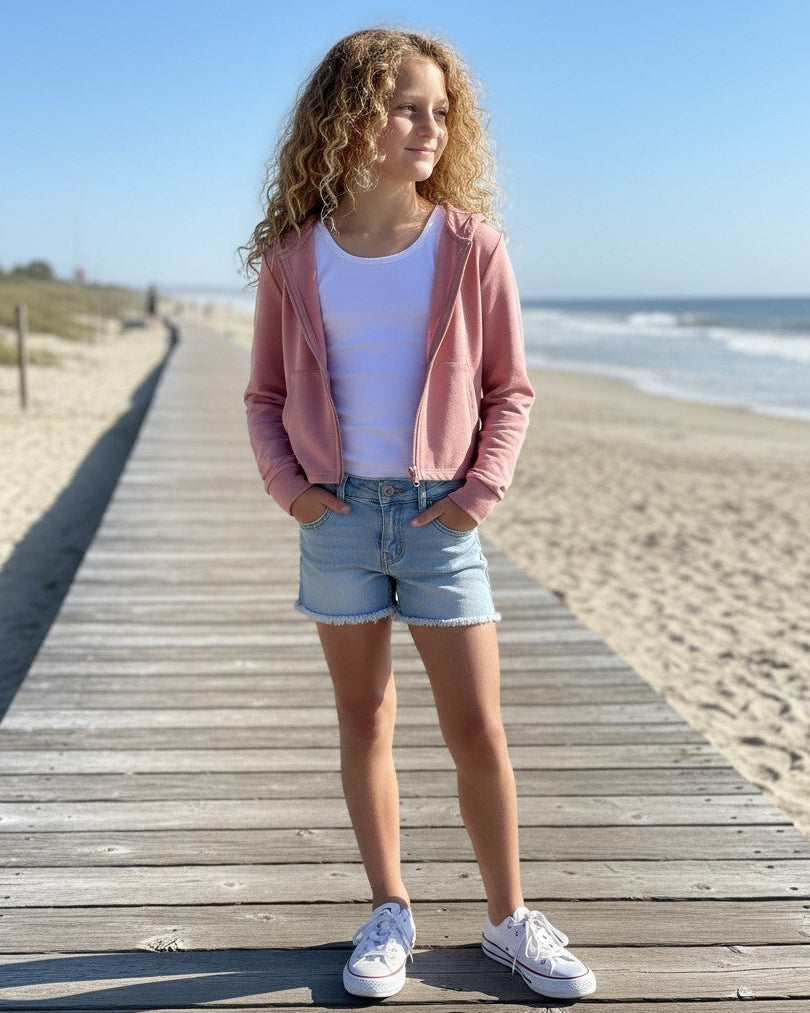 Model wearing Calla Lily Cosmetics + CL kids pink French Terry crop zip-up hoodie on a wooden beach boardwalk.
